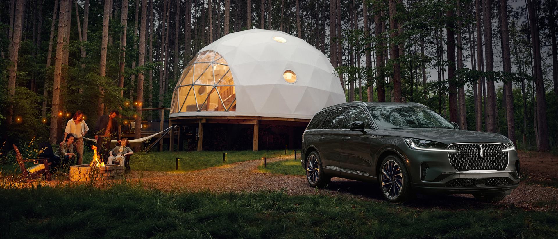 A 2027 Lincoln Aviator® SUV is parked next to a luxury camping site as a family enjoys a campfire together
