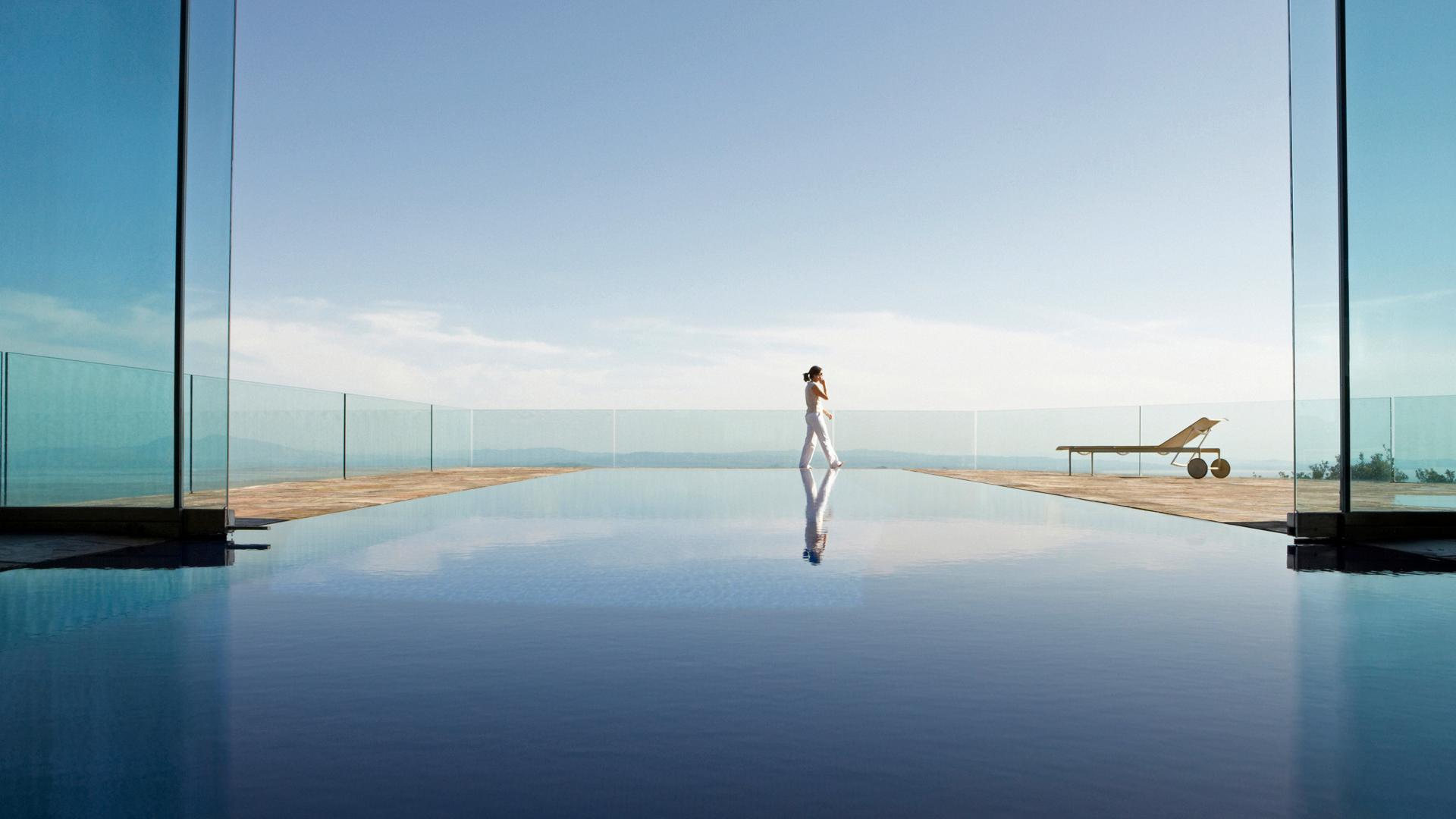 Una mujer camina junto a una piscina infinita con espejos enmarcada por paredes de vidrio, con un vasto cielo y un paisaje distante más allá