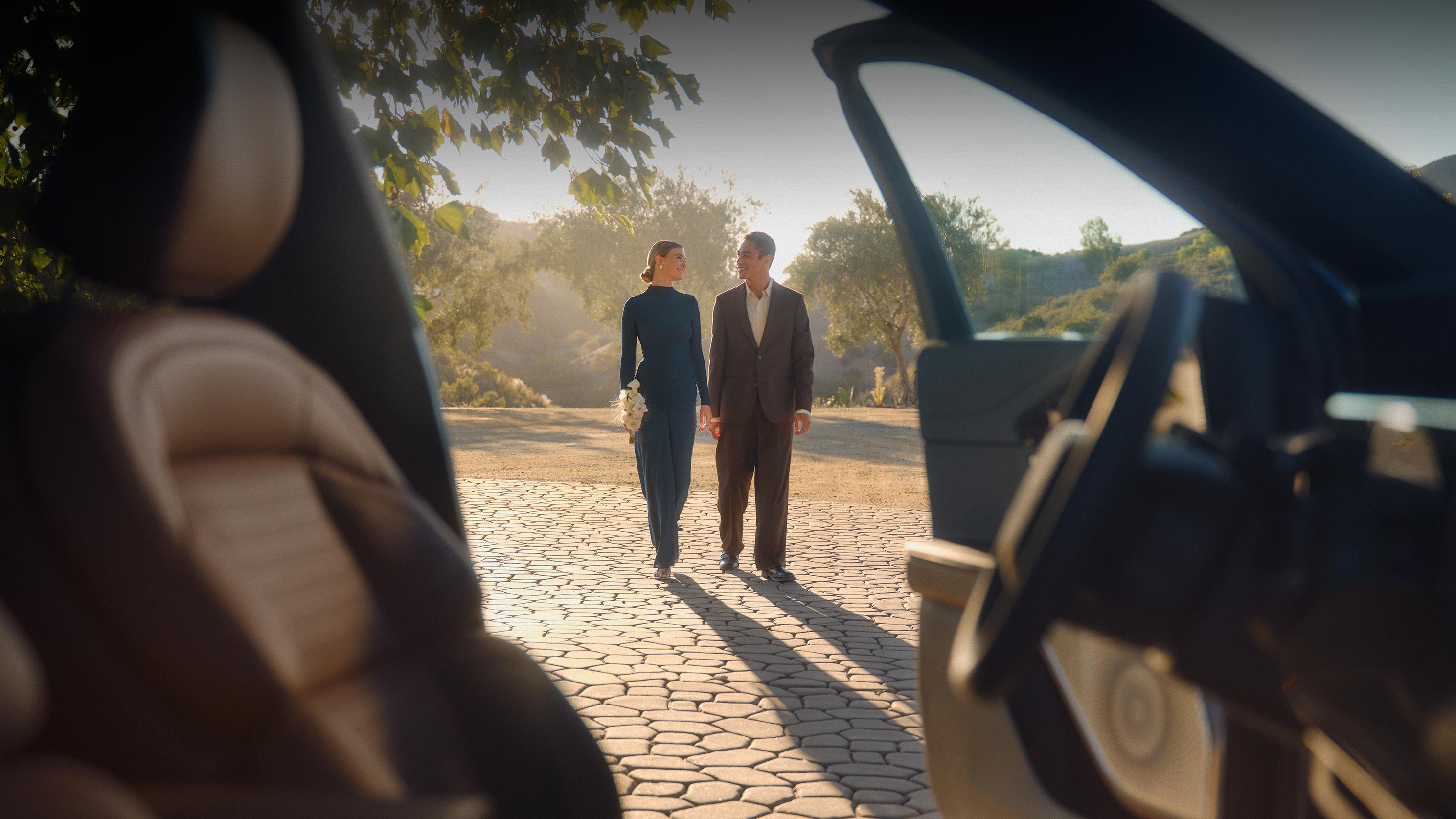 A man and a woman holding hands while looking at eachother outside a Lincoln vehicle.
