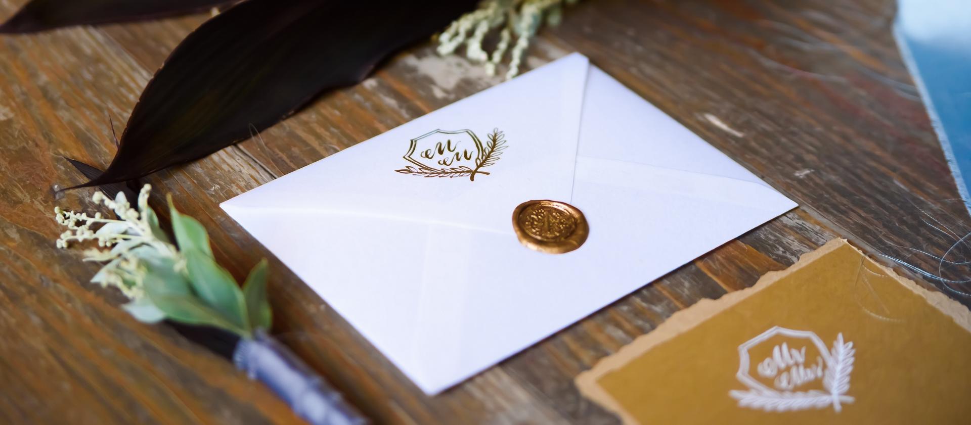 Un sobre con un sello decorativo que contiene una invitación yace sobre una superficie de madera con arreglos florales