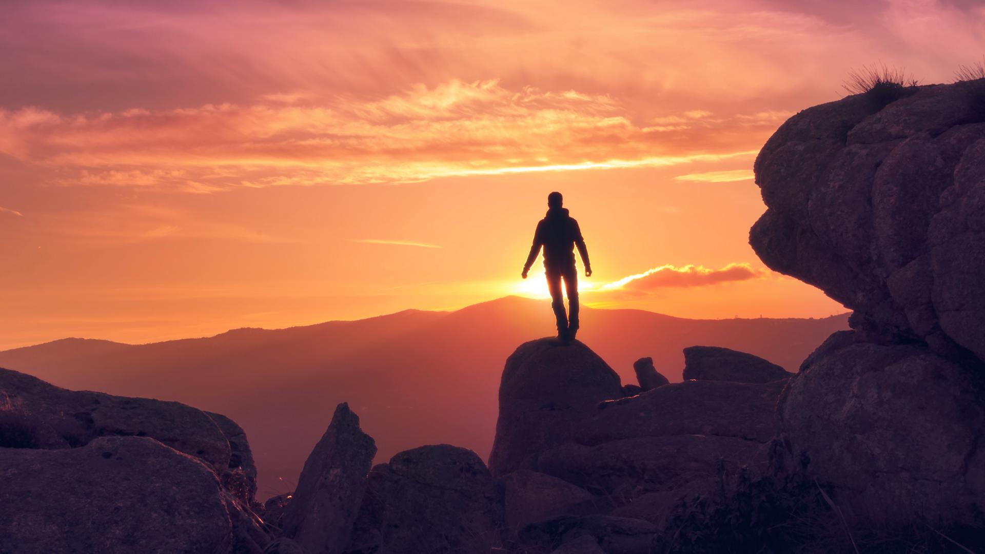 Una silueta se encuentra en la cima de una cumbre rocosa, retroiluminada por una puesta de sol dorada sobre un paisaje montañoso