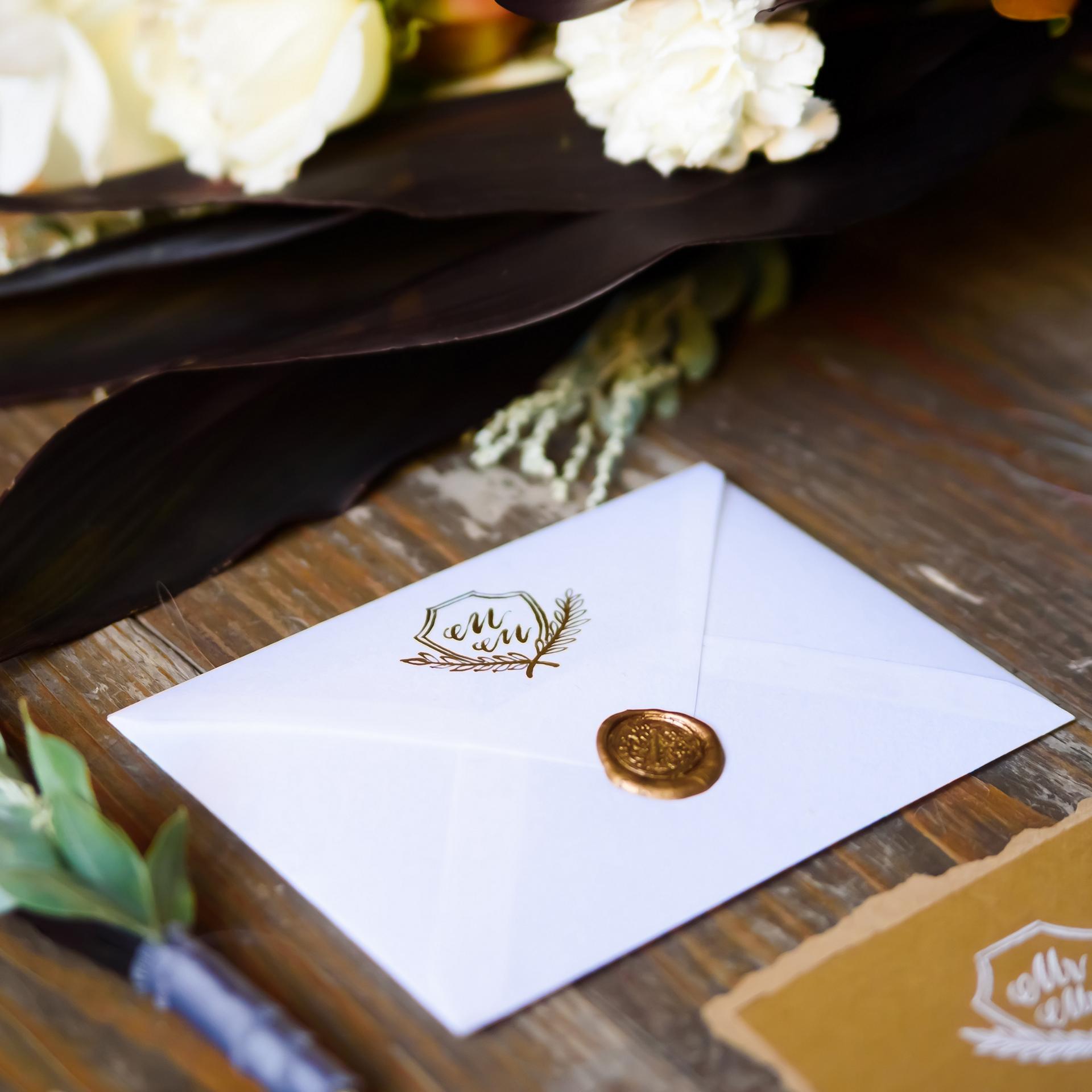 An envelope with a decorative seal holding an invitation lays on a wooden surface with floral arrangements