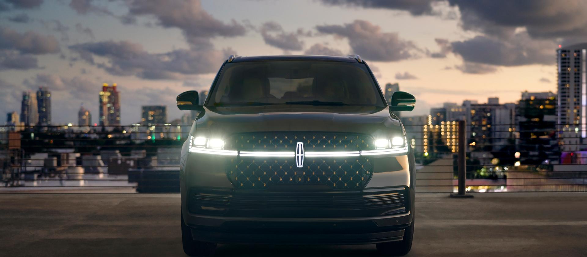 La parte delantera de una Lincoln Navigator® 2026 tiene sus faros delanteros y la Estrella Lincoln iluminados contra un telón de fondo de ciudad al anochecer