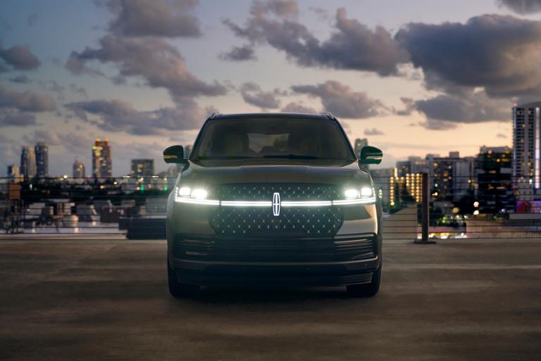 La parte delantera de una Lincoln Navigator® 2026 tiene sus faros delanteros y la Estrella Lincoln iluminados contra un telón de fondo de ciudad al anochecer