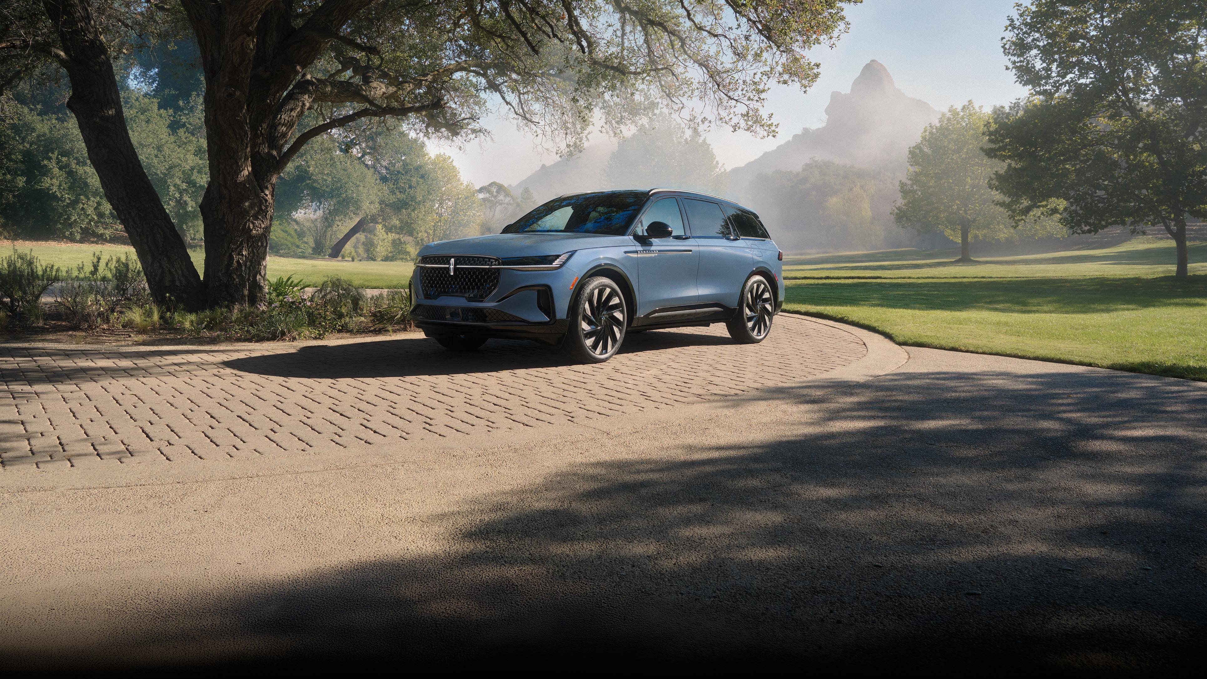 A blue 2026 Lincoln Nautilus SUV is parked on a paved path surrounded by green grass and trees